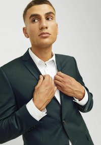 Sötétzöld öltönyzakó textúrázott felülettel, fehér ing fölé megviselve. A modell a zakó hajtókáját igazítja, bemutatva a gombokat és a mandzsetta részleteit.