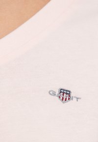 Világos rózsaszín pamut V-nyakú póló, melyen kis szürke és piros hímzett logó látható, pajzs alakú díszítéssel és csillagmotívumokkal.