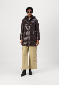 Bruine glanzende pufferjas met een capuchon, ritssluiting en zijzakken, gecombineerd met beige cargobroek en zwarte enkellaarzen.