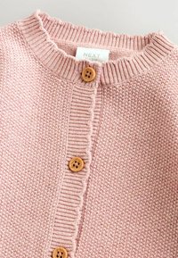 Rosa gestrickte Strickjacke mit einem strukturierten Muster, geripptem Halsausschnitt und Holzknöpfen entlang der Vorderseite, mit einem gewellten Abschluss.