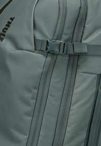 Sac à dos vert foncé avec un extérieur en textile, plusieurs compartiments zippés et une sangle avec boucle en plastique robuste.
