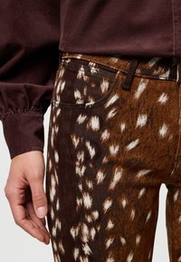 Jeans marroni e bianchi con motivo a fantasia e finitura testurizzata, dotati di una tasca frontale e di un design aderente. Abbinati a una maglia color bordeaux.