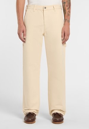 Hombre con pantalones beige de pierna ancha, zapatos mocasín de cuero marrón y camisa blanca, con el antebrazo derecho tatuado visible.