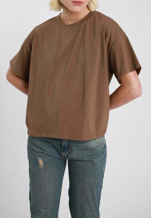 Persona che indossa una maglietta marrone a maniche corte e jeans blu sbiaditi, con le mani posizionate dietro la schiena.