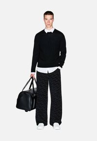 Maglione nero sopra una camicia bianca con colletto, abbinato a pantaloni neri ampi con motivo. Tiene una borsa da viaggio nera, indossa scarpe da ginnastica bianche.