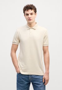 Beige pólóing rövid ujjal, klasszikus gallérral, egygombos gombolással és diszkrét logóval. Kék farmernadrággal viselve. Puha tapintás.