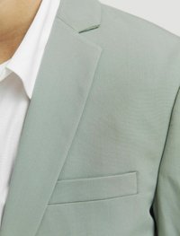 Ljusgrön blazer i ett textured tyg, med slagkrage och bröstficka, över en vit skjorta.
