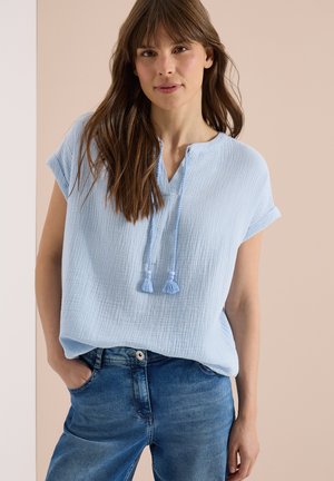 Junge Frau mit langen braunen Haaren, die ein hellblaues, strukturiertes Kurzarmoberteil mit Fransen und blaue Jeans trägt, steht mit einer Hand in der Tasche.