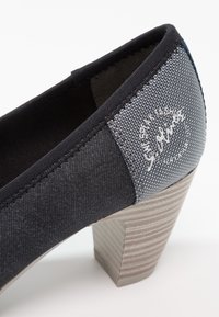 Chaussure à talon haut noire en textile avec un accent en tissu gris portant un petit logo, et un talon en bois strié. Texture globale lisse.