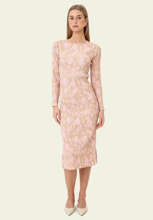 Naine, kes kannab beežikat pöidlapikkust kleiti pikkade varrukatega ja roosade lillemustritega, jalas valged terava tipuga kontsakingad, seisab ühevärvilise tausta ees.