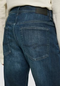 Donkerblauwe denim jeans met een gestructureerde afwerking, met achterzakken voorzien van zigzagstiksels en een zwarte leren taillebandlabel.
