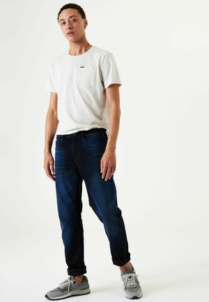 Light gray crew neck t-shirt with pocket, paired with dark blue denim jeans. Model wears gray sneakers. Casual fit, minimalistic design.