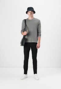 Grå långärmad skjorta, svart bucket-hatt, svarta byxor och vita sneakers. Bär en svart axelremsväska. Minimalistisk design och neutrala färger.