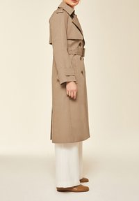 Femme portant un long trench-coat beige avec ceinture sur un pantalon large blanc et des chaussures plates marron, debout devant un fond uni.