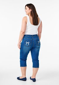 Tumši zili denim capri džinsi ar kabatām, kas rotātas ar gaiši zilu loku izšuvumu. Valkāti ar baltu bezpadušu krekliņu un tumši zilu plakanām apaviem.