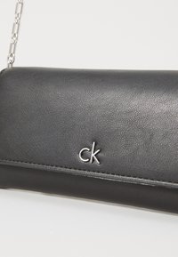 Czarna skórzana kopertówka o fakturowanej powierzchni, z srebrnym łańcuszkiem i małym metalowym logo CK z przodu.