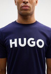 Tumši zila kokvilnas T-kreklu ar lieliem baltiem "HUGO" burtiem priekšpusē, apaļa kakla izgriezums un īsas piedurknes. Gluda tekstūra.