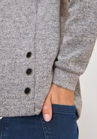 Grauer gestrickter Pullover mit subtiler Textur, ausgestattet mit drei schwarzen Knöpfen an der Seite und eng anliegenden Bündchen. Getragen über dunklen Jeans.