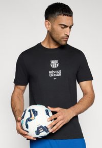 Μαύρο κοντομάνικο μπλουζάκι με το λογότυπο της Barcelona και το κείμενο "MÉS QUE UN CLUB," συνοδευόμενο από μια λευκή και μπλε μπάλα ποδοσφαίρου με σχέδιο.