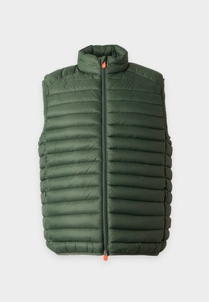Jeune homme debout les mains dans les poches, portant un gilet matelassé vert, un pull grenat, un pantalon noir et des baskets blanches sur un fond uni.