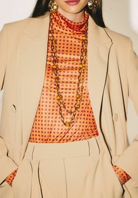 Blusa arancione a pois sotto un blazer beige, con tessuto strutturato e collana a strati con dettagli gialli e catenine.