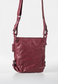 Bordeaux leren crossbody tas met een gestructureerde afwerking, verstelbare schouderriem, ritssluiting en metalen hardware accenten. Compacte rechthoekige vorm.