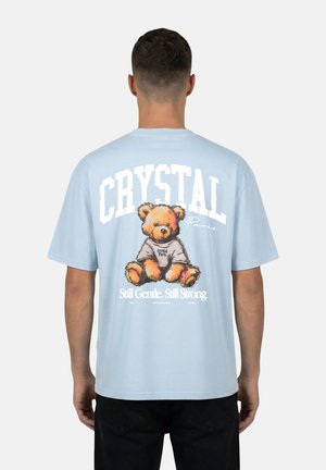 Mann von hinten gesehen, trägt ein hellblaues T-Shirt mit einem Teddybär und dem Schriftzug „CRYSTAL Paris Still Gentle. Still Strong“ auf dem Rücken.