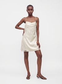 Femme portant une robe nuisette en satin beige clair avec des détails en dentelle et fines bretelles, debout avec une main sur la hanche et portant des sandales noires à talons bas.