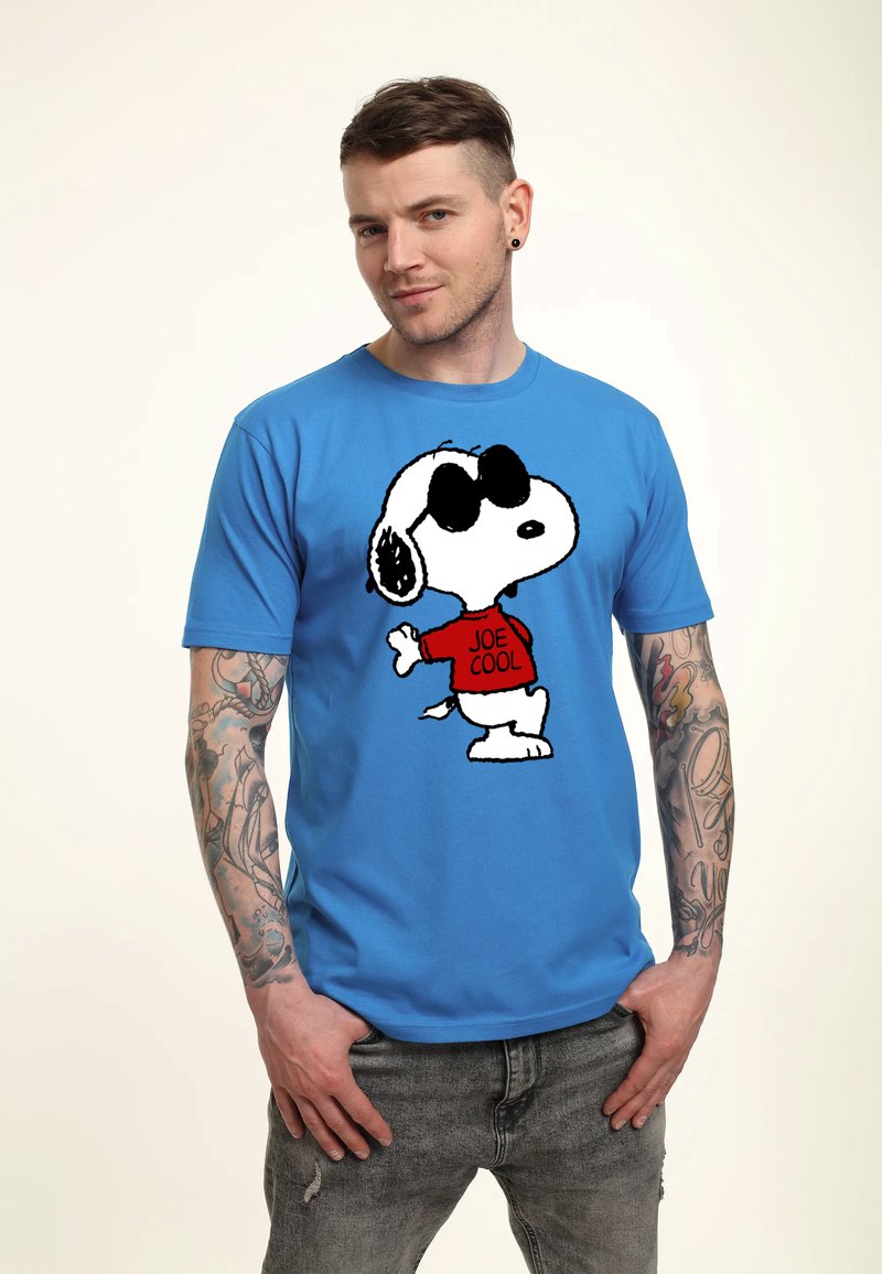 Młody mężczyzna z wytatuowanymi ramionami noszący niebieską koszulkę z postacią Snoopy w okularach przeciwsłonecznych oraz czerwoną koszulę z napisem „Joe Cool.”