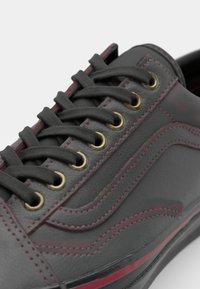 Basket en cuir noir avec accents rouges, dotée d'œillets en or, de lacets plats et d'une surface texturée. Design sur le côté avec des détails de couture.