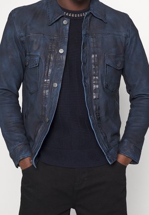 Homme portant une veste en cuir bleu marine avec des poches à rabat boutonnées sur un pull foncé tricoté, les mains dans les poches d'un pantalon noir.