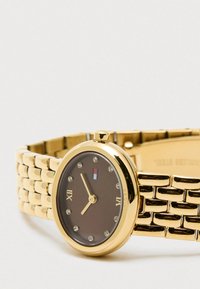 Złoty zegarek na rękę z brązową tarczą, rzymskimi cyframi przy 12 i 6, kryształowymi znacznikami godzin oraz małym logo w kolorze czerwonym, białym i niebieskim.