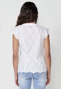 Blouse blanche brodée à manches courtes, avec des motifs floraux, des bords festonnés et une texture lisse. La vue arrière montre des coutures détaillées.
