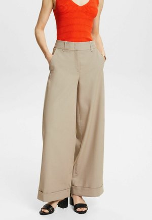 Femme portant un pantalon beige à jambes larges, un top orange sans manches et des sandales à talons noires, les mains dans les poches, sur un fond uni.