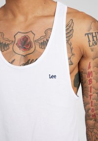Hvid bomulds tanktop med rund hals og broderet "Lee" logo i mørkeblå. Viser tatoveringer på armen og brystet.