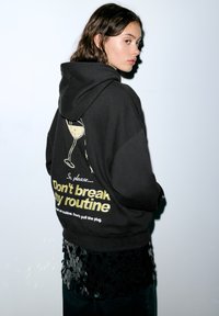 Zwarte hoodie met een afbeelding van een glas en de tekst "Don't break my routine" in goud. Zachte stof, oversized pasvorm en met capuchon.