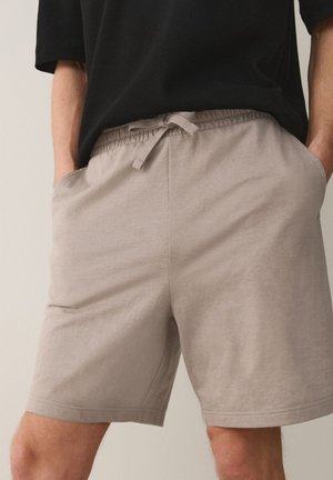 Persona che indossa pantaloncini beige con coulisse e una maglietta nera, in piedi con le mani in tasca su uno sfondo semplice.