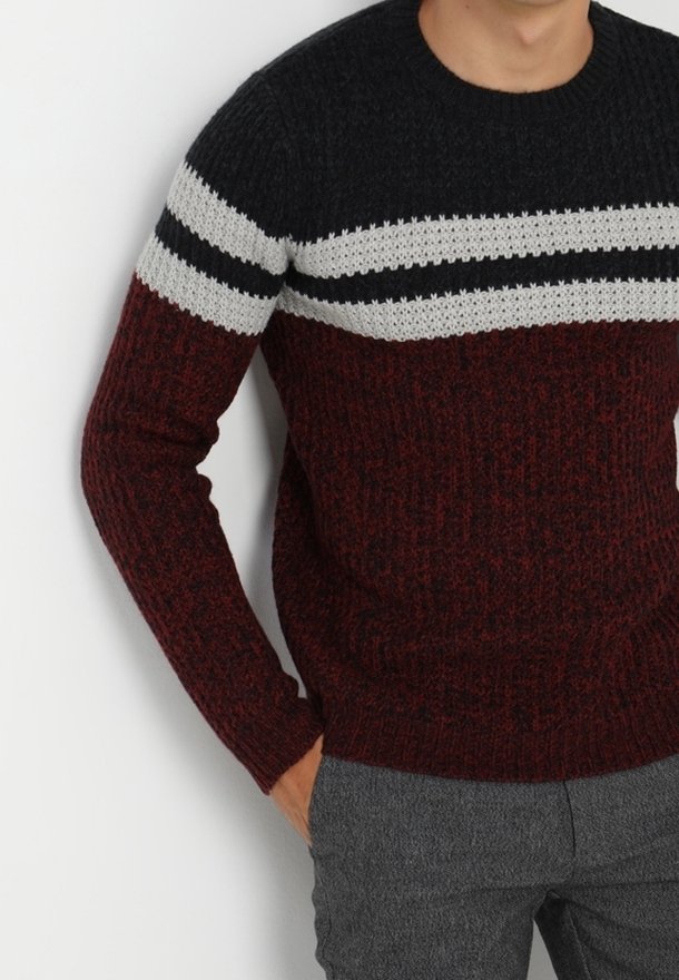 Homme portant un pull en maille bordeaux et noir avec des rayures horizontales blanches et noires sur la poitrine, assorti à un pantalon gris.