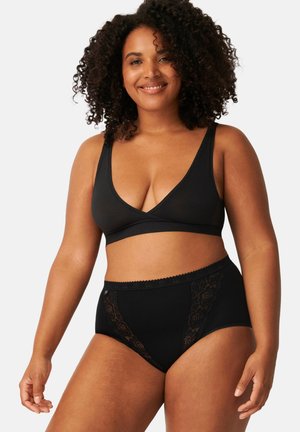 Lächelnde Frau mit lockigen Haaren, die einen schwarzen Bralette und hoch taillierte schwarze Unterwäsche mit Spitzenbesatz vor weißem Hintergrund trägt.