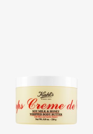 Beurre corporel fouetté dans un pot rond jaune avec un couvercle blanc. Présente un texte rouge et un logo noir. Contient du lait de soja, du miel, du beurre de karité et du jojoba.