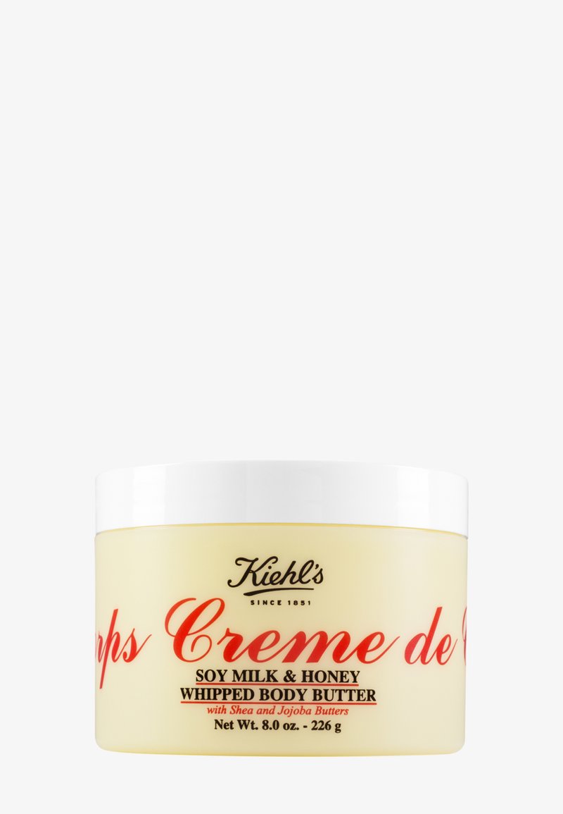 Manteca corporal batida en un tarro redondo amarillo con tapa blanca. Presenta texto rojo y un logo negro. Contiene leche de soja, miel, manteca de karité y manteca de jojoba.