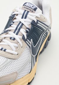 Nike Vomero 5-sneakers i grått, marinblått och beige. Har nätöverdel, snörning, gummisula och anmärkningsvärd branding på tungan och sidan.