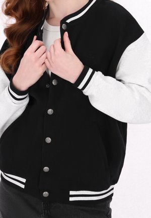 Frau trägt eine schwarz-weiße College-Jacke mit Druckknöpfen, hält den Kragen mit beiden Händen, langes rotes Haar sichtbar.