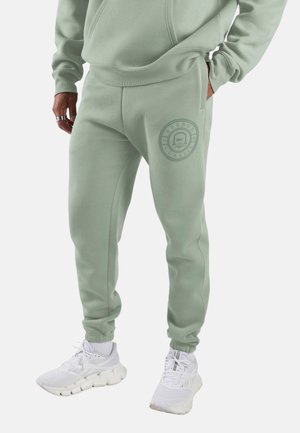 Man draagt lichtgroene Reebok Classic joggingbroek met elastische boorden en witte sportschoenen, staand met één hand in de zak.