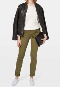 Giacca di pelle nera, maglione bianco, pantaloni slim-fit verde oliva e sneakers bianche. Tiene una pochette nera.