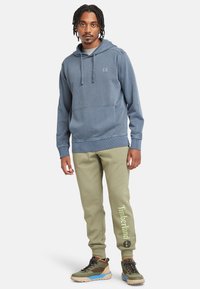 Hanorac albastru din bumbac cu buzunar frontal, tricou gri dedesubt, pantaloni de trening verde măsliniu cu logo-ul "Timberland" și pantofi sport maro.