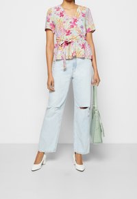 Haut floral en pastel avec des manches courtes et une taille nouée. Porté avec un jean déchiré à lavage clair et des chaussures à talons blocs blanches. Sac vert.