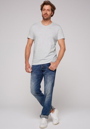 Man standing with arms relaxed, wearing a light gray short-sleeve V-neck t-shirt, blue jeans, and white sneakers against a plain background.