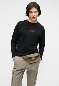 Νεαρό άτομο με σκουρόχρωμα μαλλιά μεσαίου μήκους φοράει μαύρο φούτερ με κόκκινο κείμενο «REPLAY» και μπεζ παντελόνι cargo με πράσινο τσαντάκι μέσης.