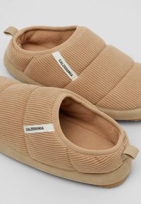 Beige quiltede hjemmesko lavet af blødt stof og fleece, med en polstret indersål, flad gummisål og en løkke ved hælen.